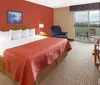 The image shows a neatly arranged hotel room with a king-sized bed a red accent wall contemporary furnishings and a view outside through a glass door