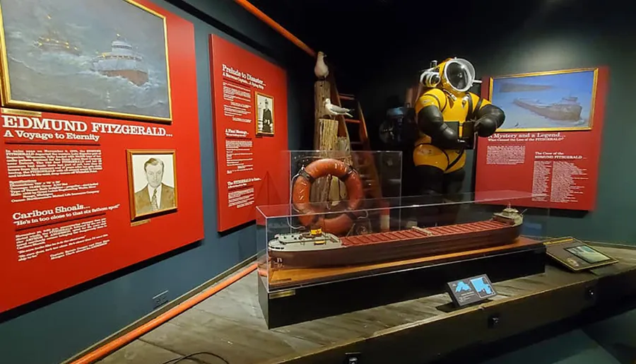 The image shows an exhibition about the ship Edmund Fitzgerald, with informational displays, a model of the ship, and a diving suit, highlighting its historical significance and maritime legacy.