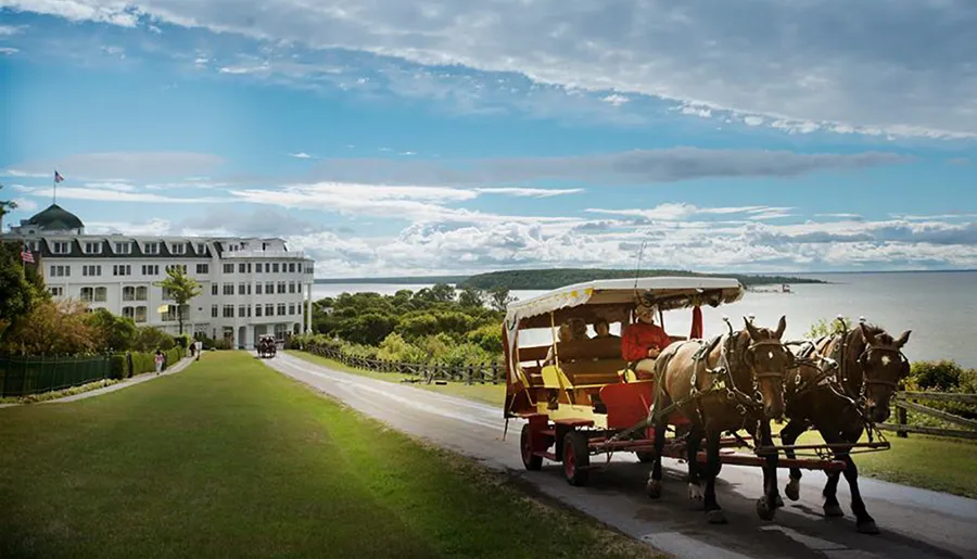 A horse-drawn carriage with passengers travels along a road near an expansive white hotel with views of a lake and green surroundings under a partly cloudy sky.