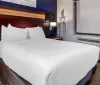 The image shows a neatly made bed in a modern hotel room with simplistic decor and a wall-mounted air conditioning unit