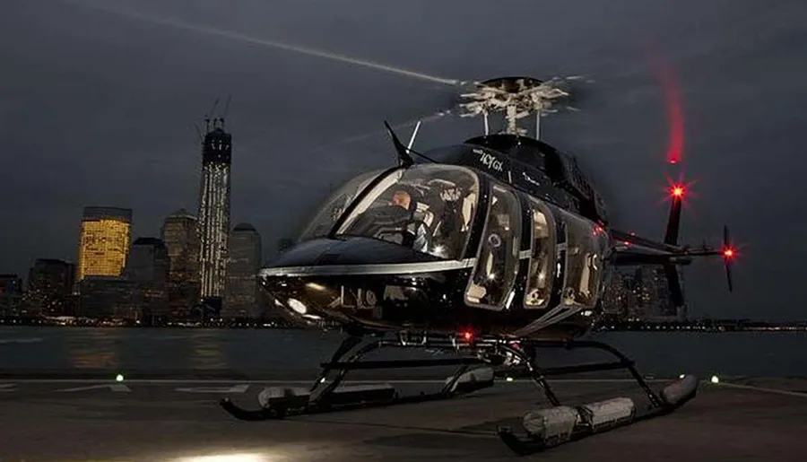 A helicopter with illuminated cabin lights is parked on a helipad at dusk with a city skyline in the background.