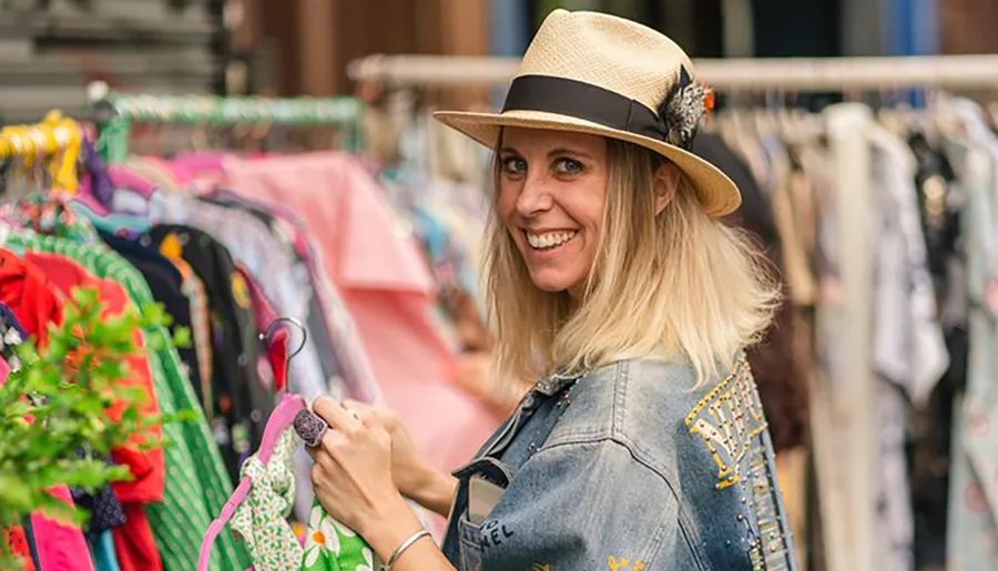 A smiling woman in a straw hat and denim jacket is browsing through clothes at an outdoor market.