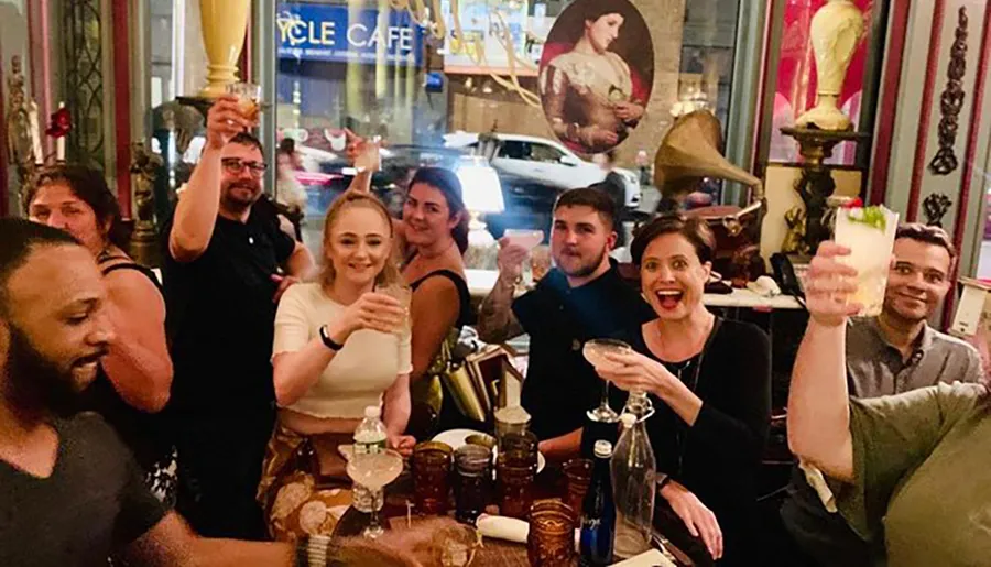 A group of people is joyfully toasting with drinks in a lively indoor setting, with a glimpse of the city street visible through the window.