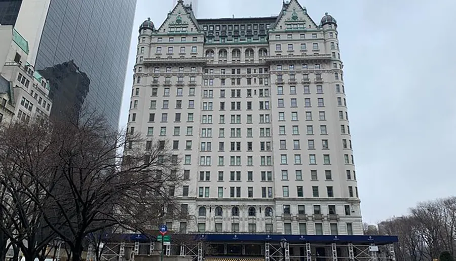 The image shows a large, historic hotel with a chateau-style roofline juxtaposed against the backdrop of modern skyscrapers on an overcast day.
