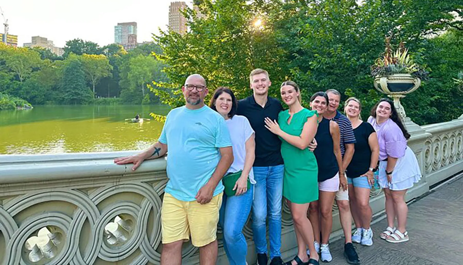 A group of eight people is posing for a smiling photo on a bridge over a pond in a lush park environment with city buildings in the background.