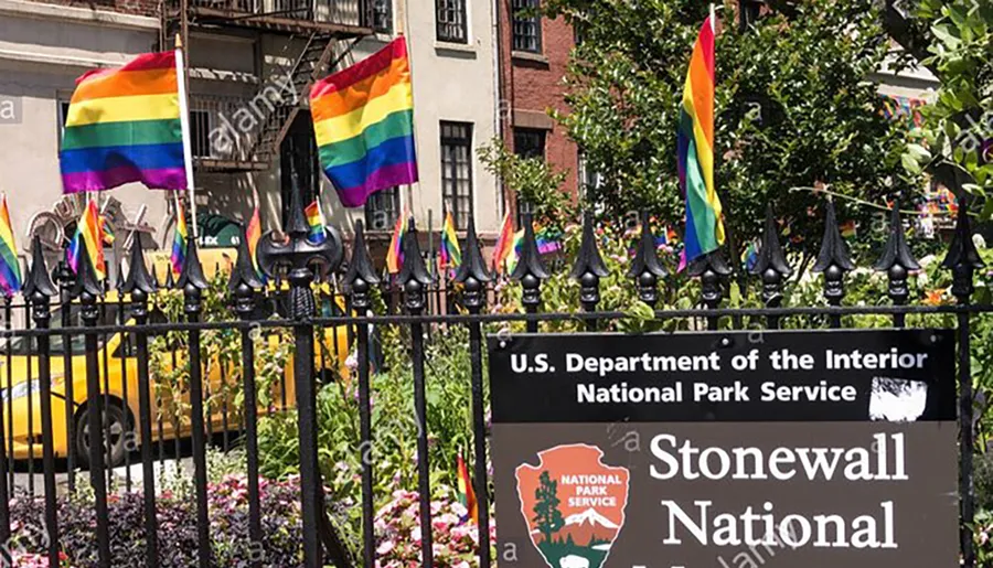 The image shows rainbow flags prominently displayed on a fence in front of a sign for the Stonewall National Monument, symbolizing LGBTQ+ pride and history.