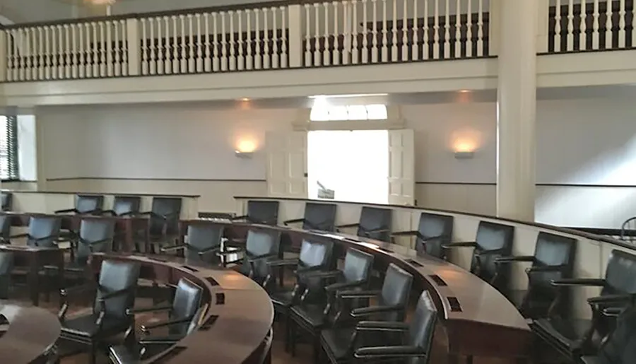 The image shows a formal meeting room with rows of upholstered chairs arranged in a semicircular pattern facing a central area, with a balcony level above.