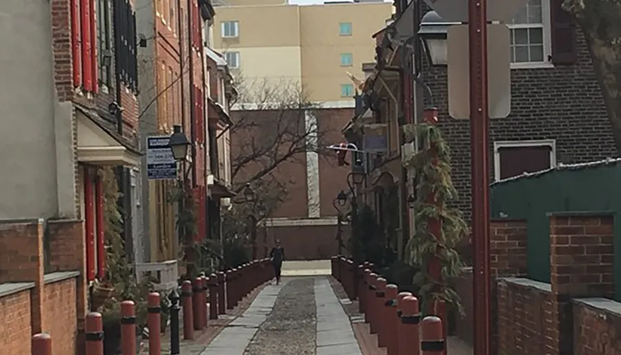 The image shows a quaint, narrow alley lined with red bollards and historical buildings, with a person in the distance walking away from the viewer.