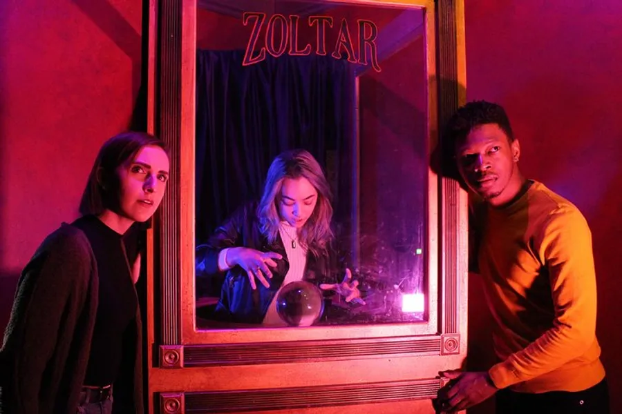 Three individuals are gathered around a Zoltar fortune-telling machine with expressions of intrigue and amusement under ambient red and purple lighting.