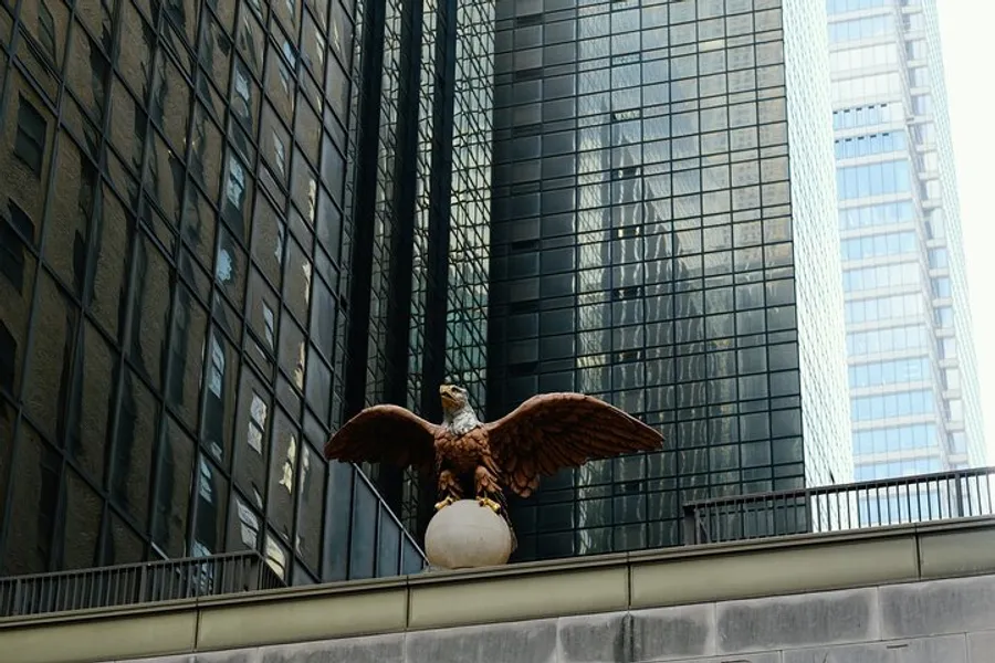 An eagle sculpture with outstretched wings perched on a sphere is set against the backdrop of modern high-rise buildings with reflective glass facades.