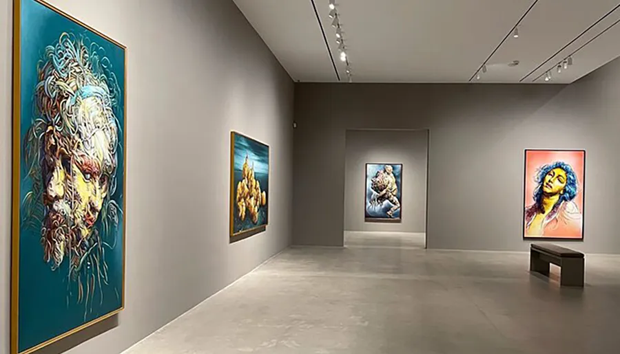 The image shows a modern art gallery with colorful, expressive paintings on the walls and a bench for viewers to sit and observe the artwork.