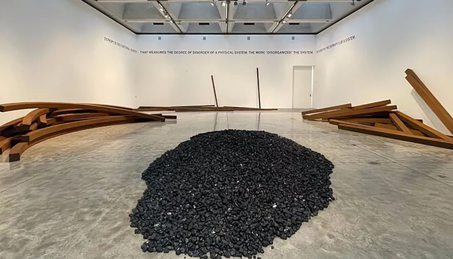 The image shows an art installation with a heap of coal at its center flanked by curved wooden structures within a gallery space, accompanied by a text on the wall.