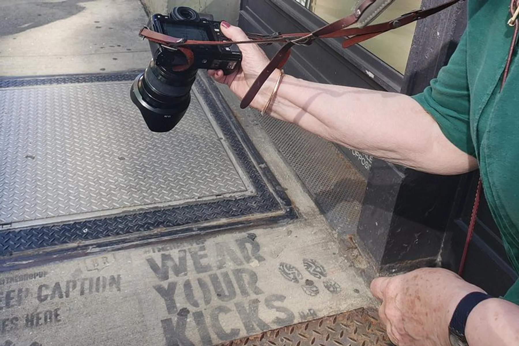A person is holding a camera next to a dirty doormat with the phrase Wear your kicks stenciled on it.