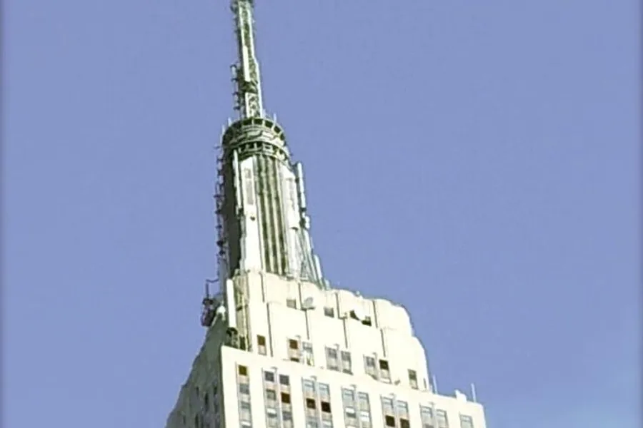 The image shows the top portion of the Empire State Building against a clear blue sky.