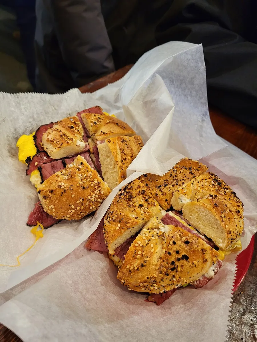 The image showcases a sliced bagel sandwich with beef and mustard on a piece of wax paper, with a person partly visible in the background.