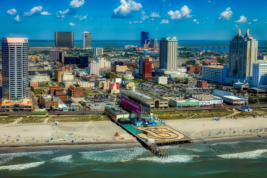 This image shows an aerial view of a vibrant coastal city with a sandy beach in the foreground, a pier extending into the sea, and a downtown skyline under a clear blue sky.
