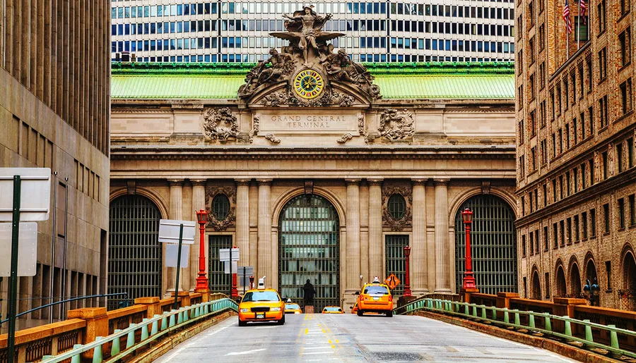 The image shows a street view of the iconic Grand Central Terminal in New York City, with yellow taxis in the foreground and the terminal's grand facade and ornate statuary clearly visible.