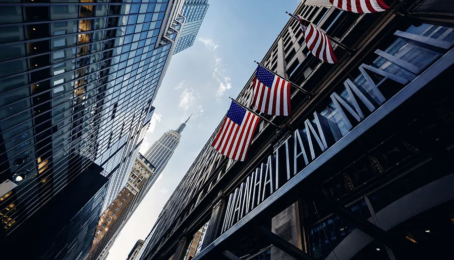 This image captures a view of urban architecture and the Empire State Building framed by American flags flying outside a building with MANHATTAN signage.