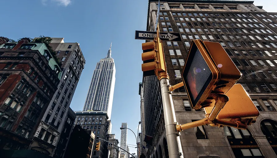 A street view featuring a traffic light and one-way sign with the Empire State Building towering in the background against a clear blue sky.