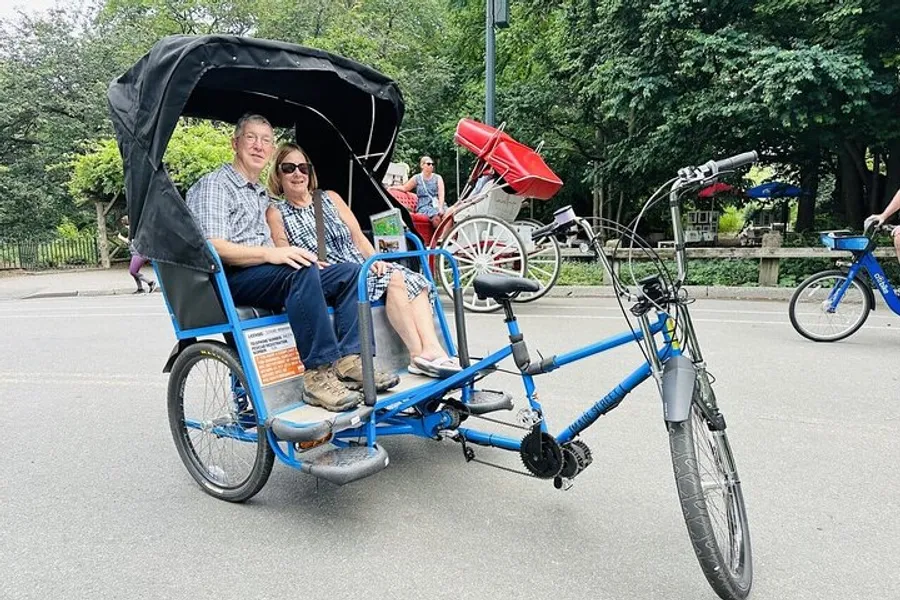 Two people are enjoying a ride on a blue pedicab through a park while another individual bikes in the background.