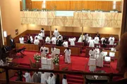 The image shows a congregation, most wearing white robes, gathered inside a church with wooden pews and a pulpit, indicating a religious ceremony or service in progress.