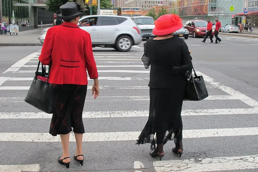 Two elegantly dressed individuals in red and black attire with matching hats are waiting to cross a bustling city street.
