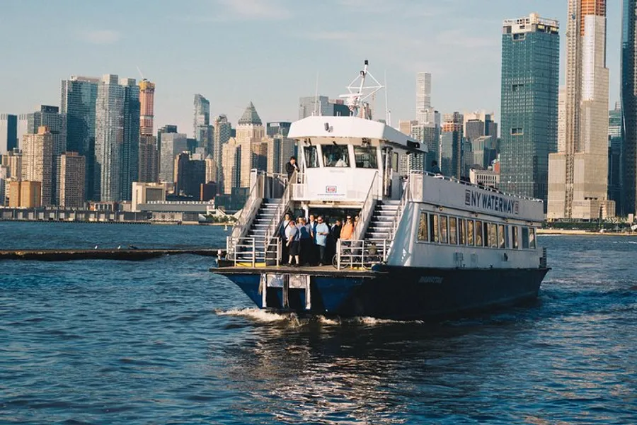 A ferry marked NY Waterway is on the water with passengers standing on the deck against the backdrop of a city skyline.