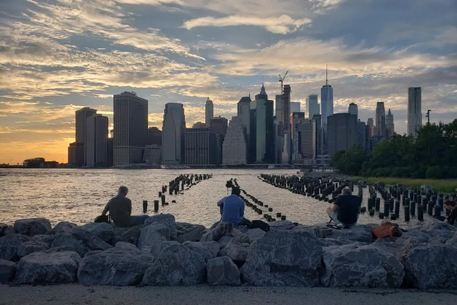 The image shows people sitting on rocks enjoying the sunset with a view of a city skyline across the water, featuring tall buildings and a tranquil atmosphere.