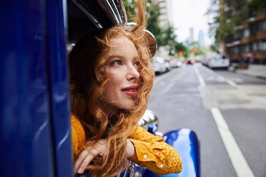 A woman with red hair is gazing out of the passenger window of a blue vehicle with a thoughtful expression, on a tree-lined city street.