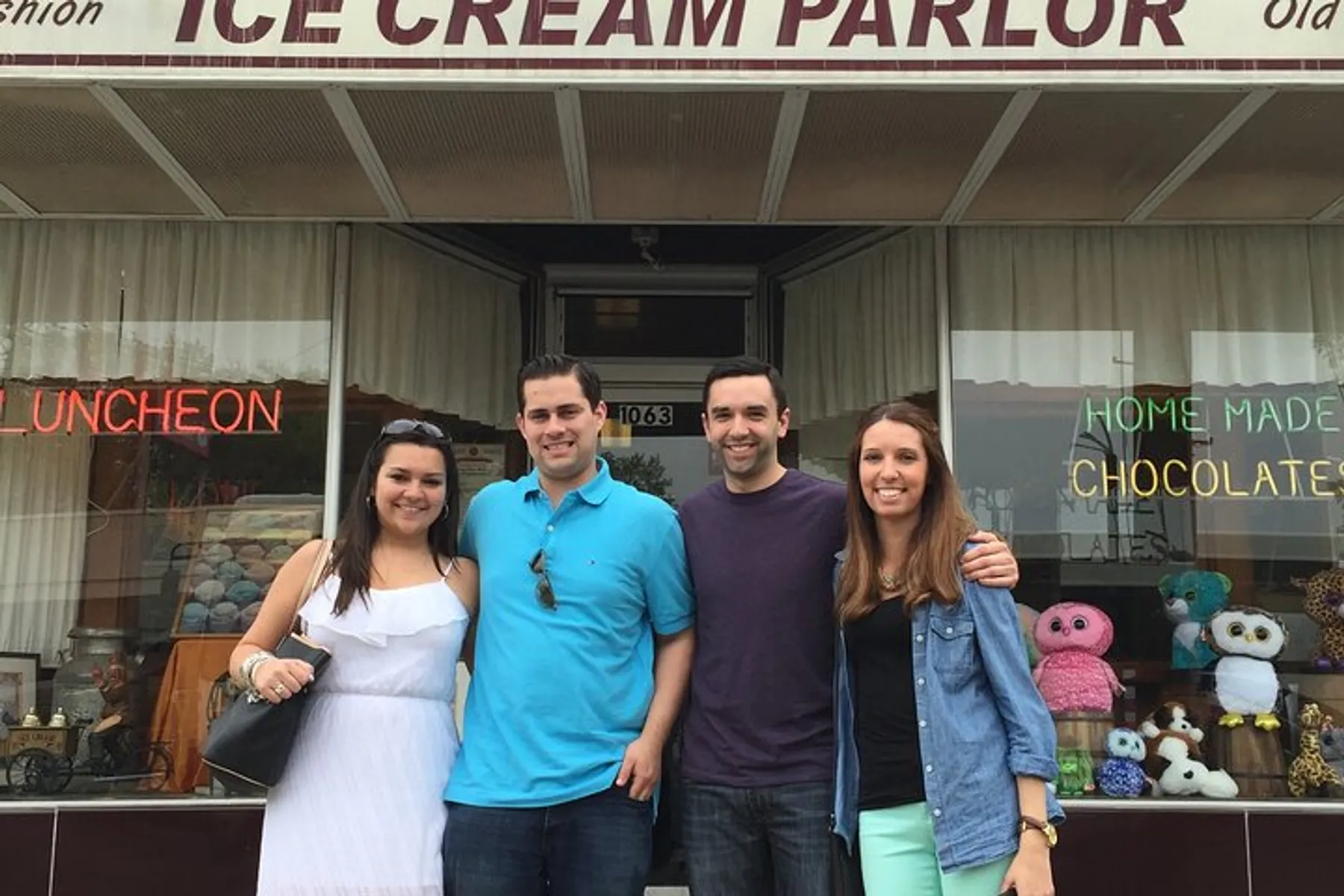 Four people are smiling in front of an old-fashioned ice cream parlor that also advertises homemade chocolates and luncheon services.