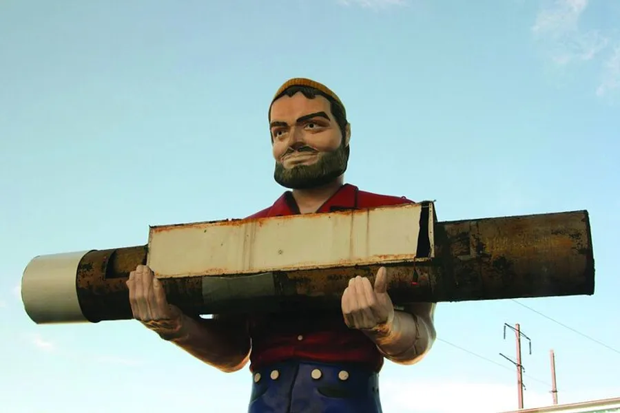 A large, colorful statue of a bearded man in a cap and rolled-up sleeves carries an oversized, cylindrical object on his shoulder against a sky backdrop.
