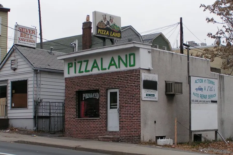 The image shows a small, brick building with a prominent green PIZZALAND sign, located on a street corner with adjacent businesses and signage.