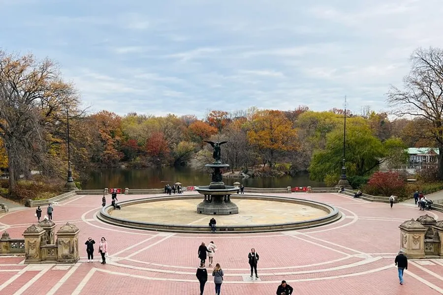 The image shows people walking around a large fountain with an angel statue on top, set within an urban park featuring vibrant autumn foliage on the trees in the background.