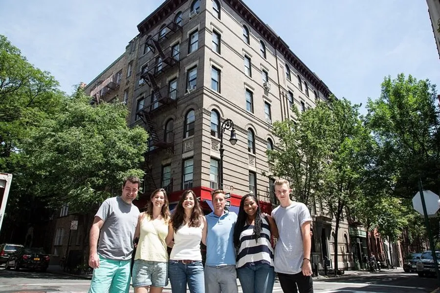 A group of seven individuals is smiling for the camera in front of an old corner building on a sunny day with trees and parked cars along the street.