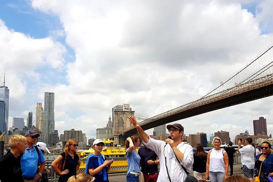 A tour guide is gesturing and explaining something to a group of attentive tourists with the Brooklyn Bridge and the Manhattan skyline in the background.