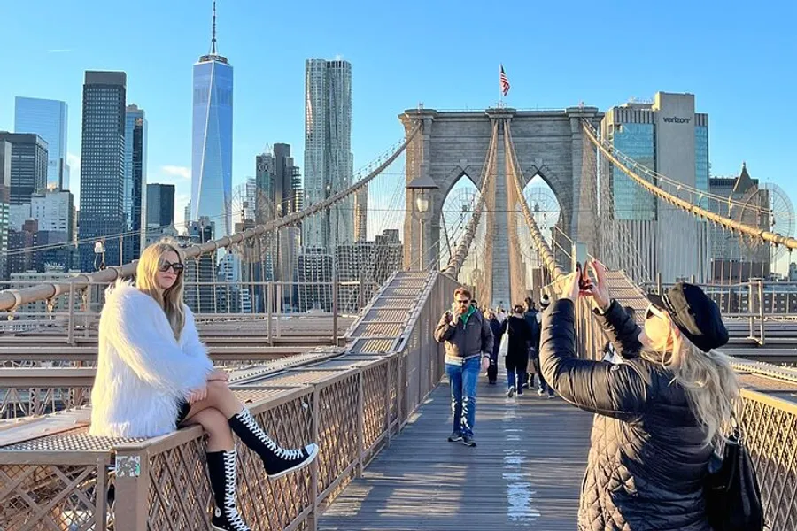 A person poses for a photo while another takes their picture on Brooklyn Bridge with the New York City skyline in the background.