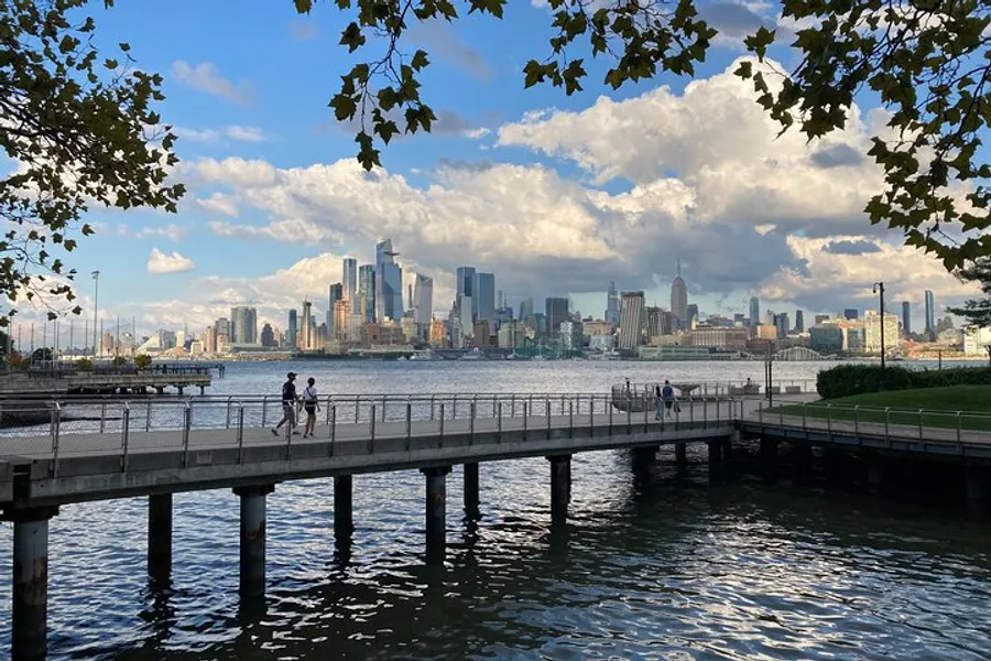 The image features a scenic view of a city skyline beyond a water body, captured from a shaded park area with people walking on a pier.