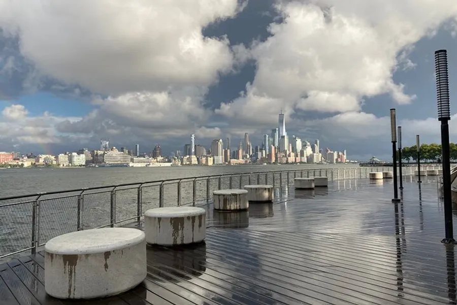 The image shows a waterfront view of a city skyline with high-rise buildings, under a dramatic sky with a partial rainbow, taken from a wet wooden pier with seating bollards.