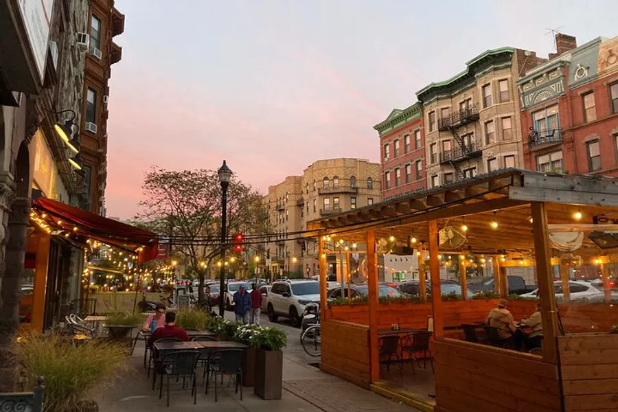 An urban street scene at dusk with outdoor dining, string lights, and vintage buildings under a pink-tinged sky.