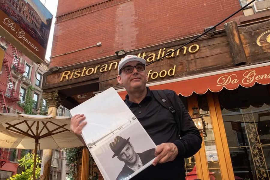 A man is standing in front of an Italian restaurant holding up a clear plastic sleeve with a black and white photograph inside.