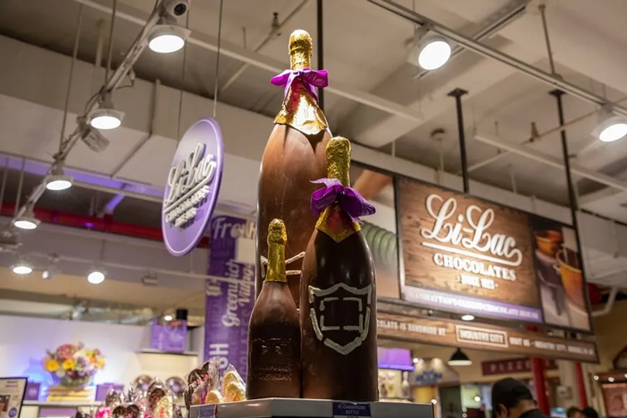 The image shows a large chocolate sculpture of a champagne bottle with a purple ribbon, displayed in a store with other chocolate products and signage in the background.