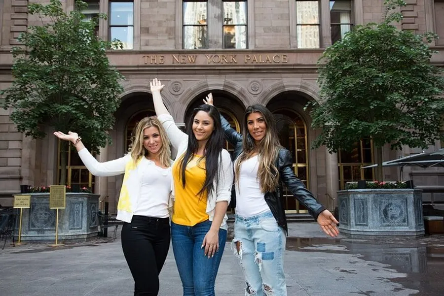 Three smiling women pose in front of the New York Palace Hotel, with one arm extended upwards.