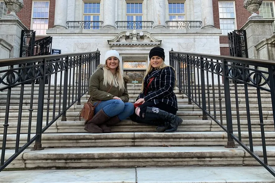 Two smiling individuals are sitting on the steps in front of an ornate building with classical architecture.