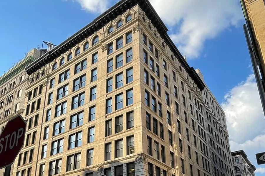 The image shows a large, multi-story corner building with classic architectural details under a blue sky with a stop sign in the foreground.