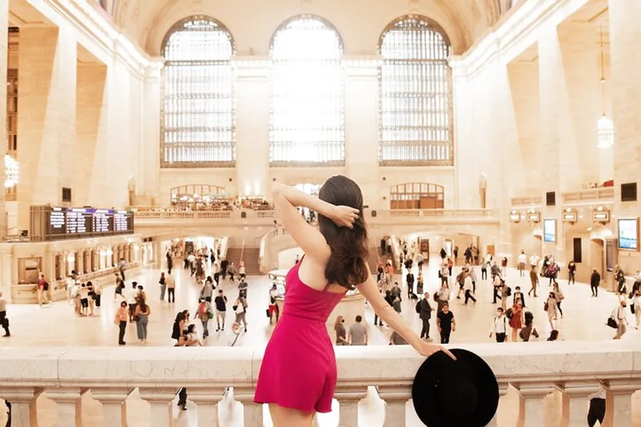A woman in a pink dress is seen from behind, holding a hat and looking over the bustling interior of a grand train station.