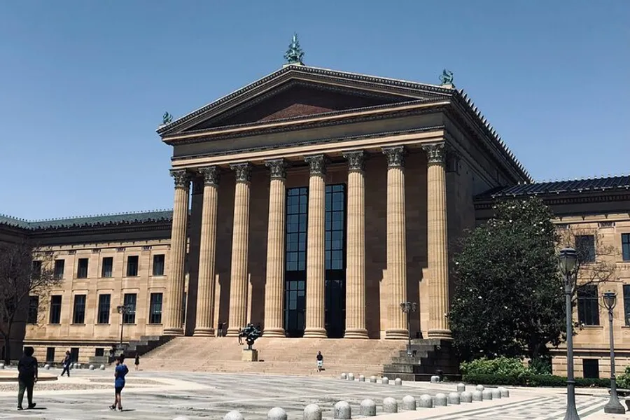 The image shows a grand neoclassical building with tall columns and a wide staircase, under a clear blue sky, with people walking in front.