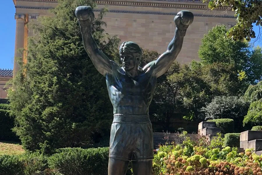 The image features a bronze statue of a boxer with his arms raised in victory, positioned in front of a building with classical architecture amidst greenery.