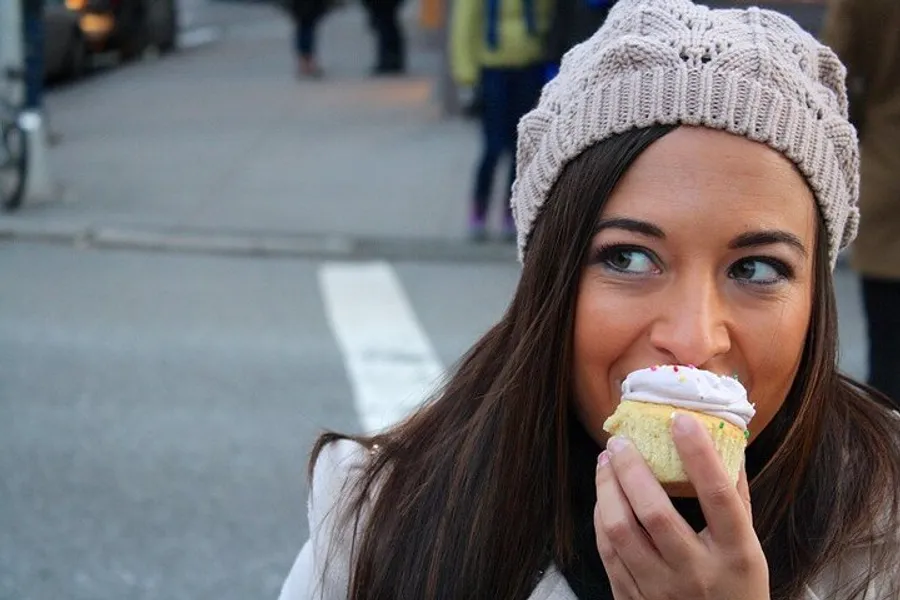 A woman wearing a knitted beanie is smiling as she is about to take a bite from a frosted cupcake outdoors.