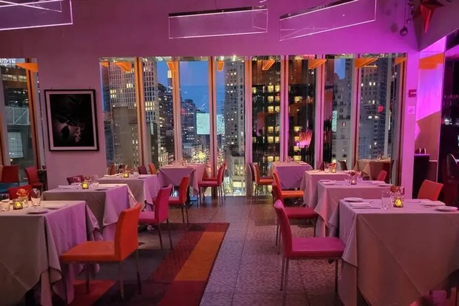 The image shows an upscale restaurant interior at twilight with neatly arranged tables and a panoramic view of a brightly-lit cityscape.