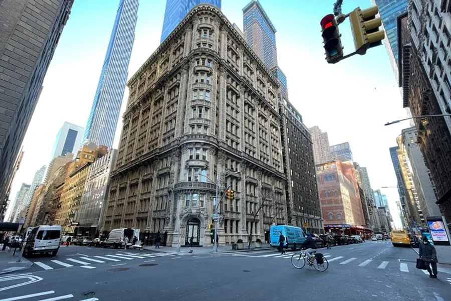 The image shows a historical, ornate building at a street corner in an urban setting, contrasted by surrounding modern skyscrapers under a bright blue sky.